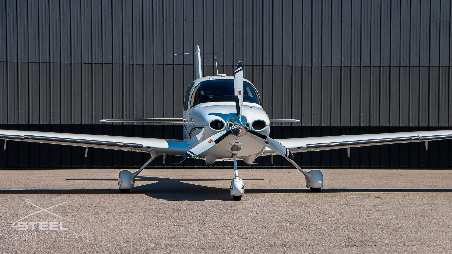 Close-up front view of a white Cirrus SR22 aircraft, highlighting its propeller and landing gear, in front of a grey hangar.
