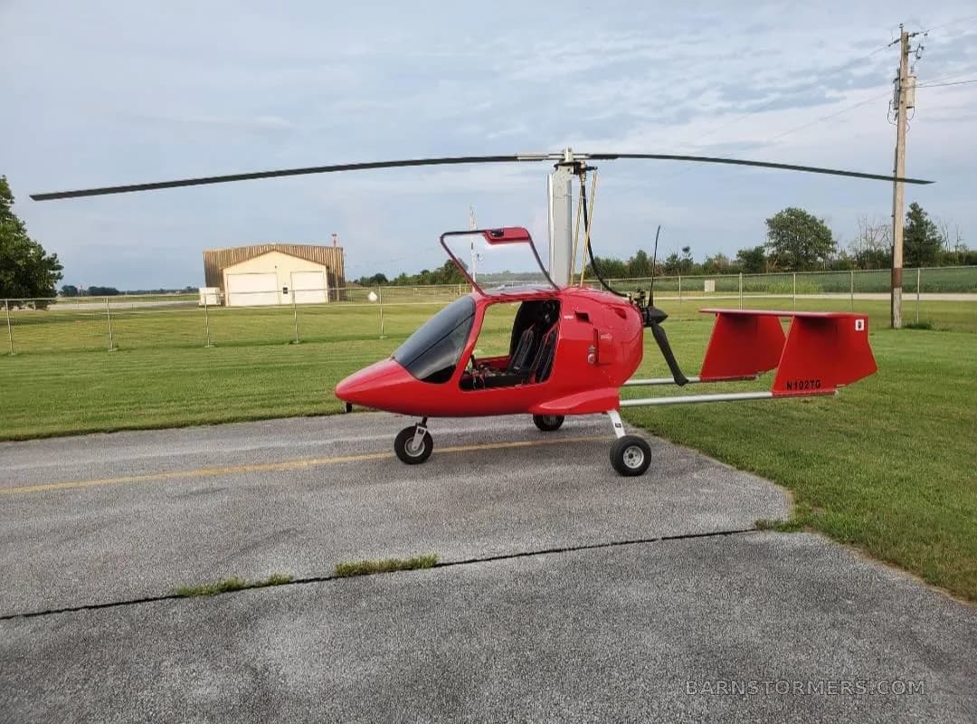 A red autogyro with its canopy open is parked on an asphalt runway next to a grassy field with hangars in the distance.