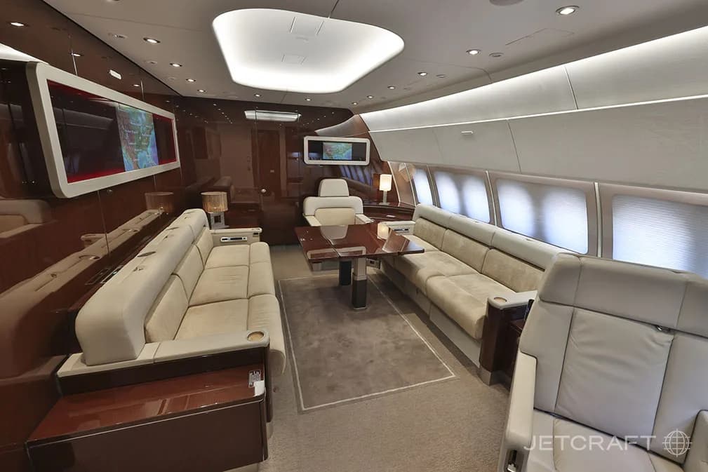 A spacious aircraft interior features two long cream-colored sofas facing each other across a dining table, with large screens on the walls.