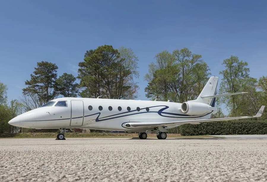 A white business jet with a distinctive blue wavy stripe on its fuselage, parked on an airfield under a clear blue sky.