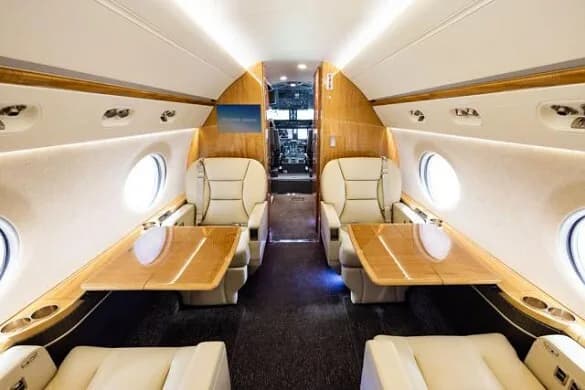 The private jet cabin interior with beige seats and wooden tables, looking towards the cockpit.