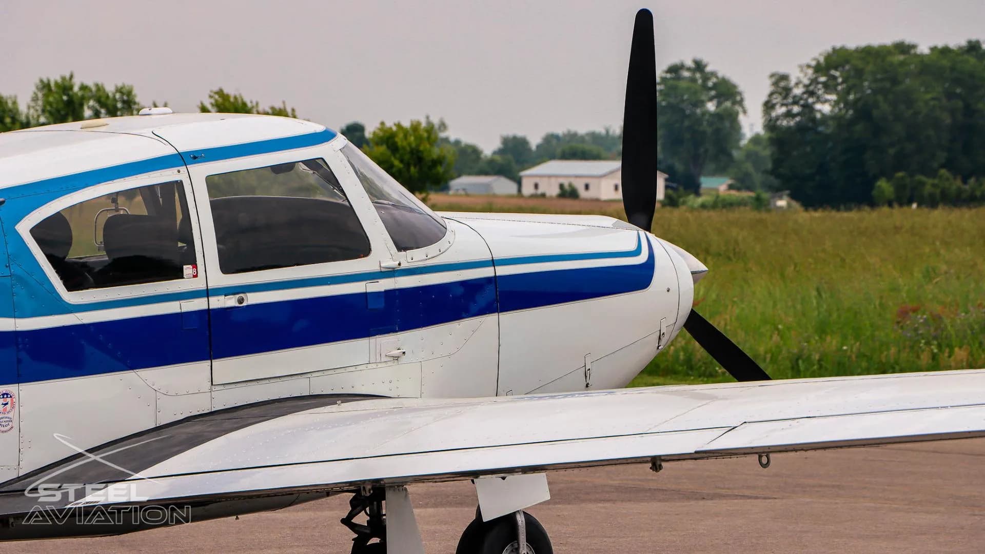 A close-up side view of the front fuselage and wing of a white and blue single-engine aircraft on a tarmac.