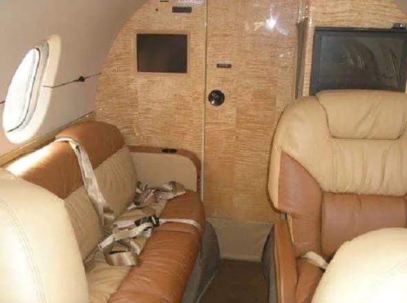 A close-up view of the private jet interior, showing a tan and brown leather seat with a seatbelt and a wooden bulkhead.