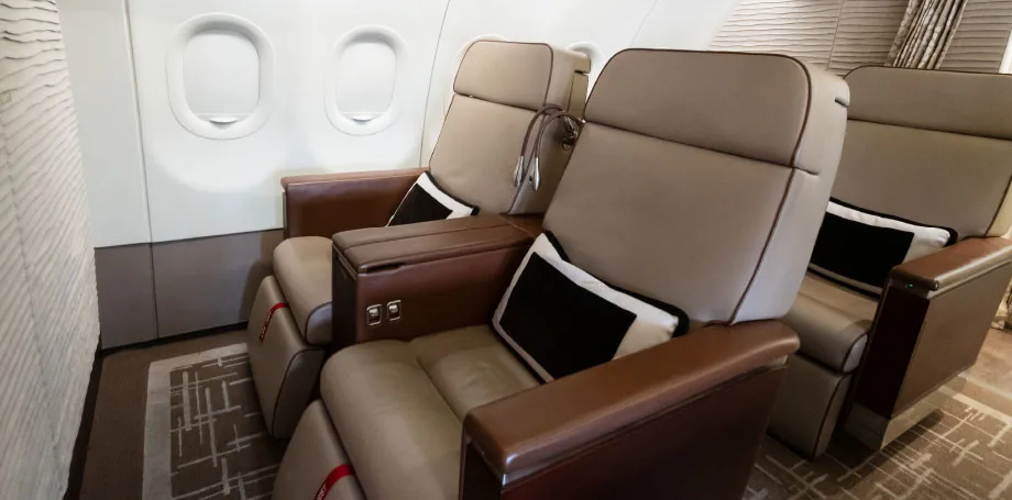 Two rows of luxurious tan leather aircraft seats with black and white throw pillows are visible next to windows.