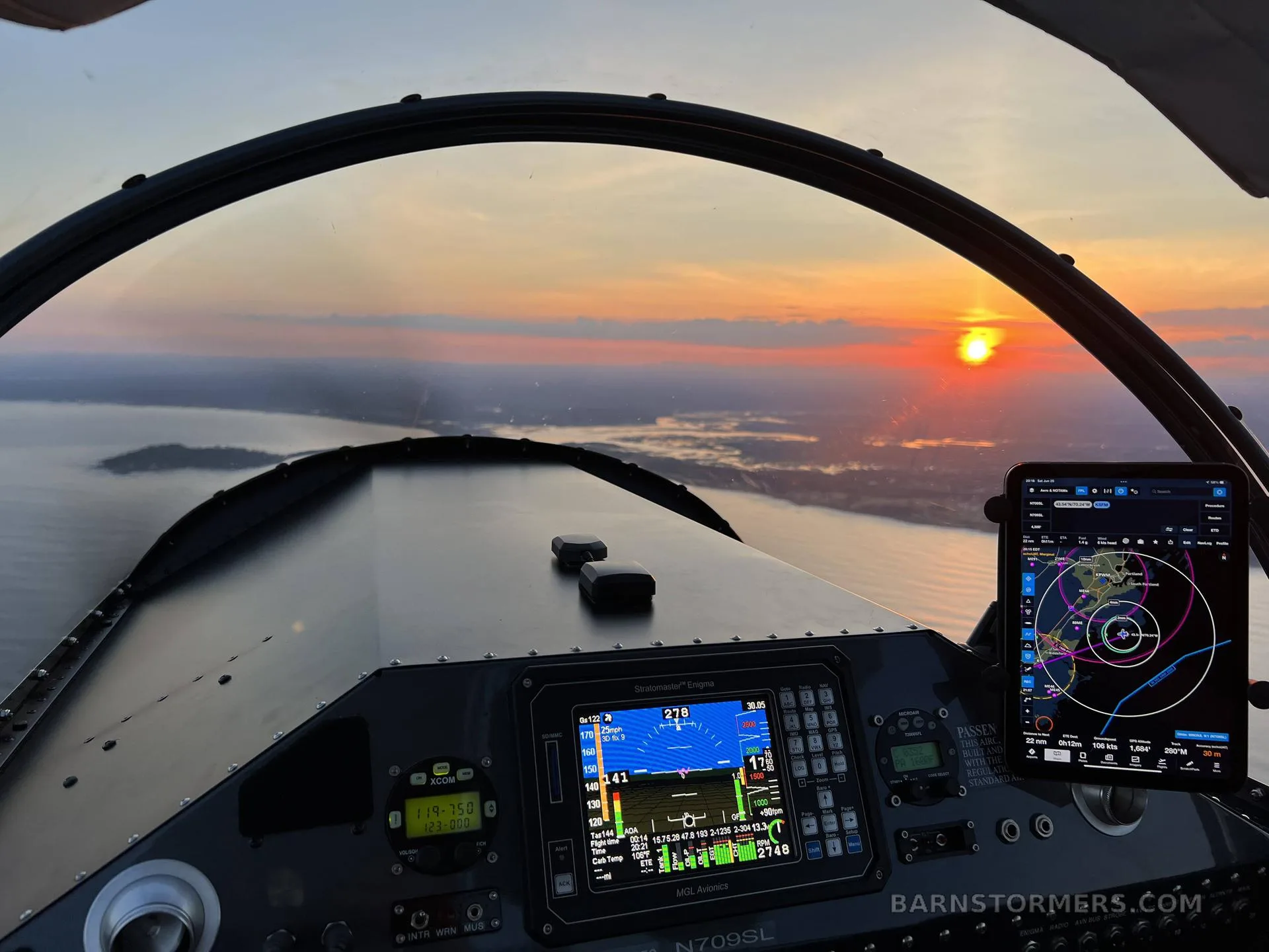 The cockpit view of an aircraft shows the instrument panel, a tablet with a map, and a sunset over water.