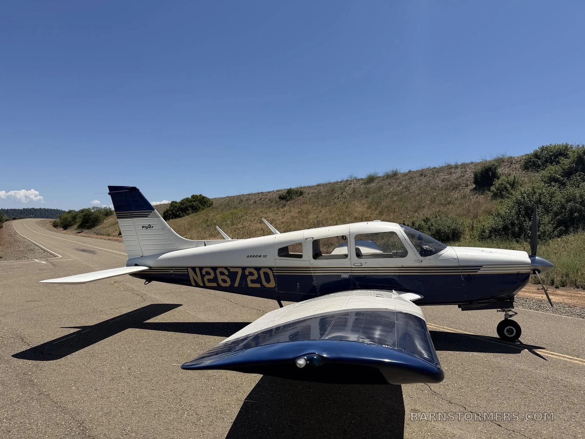 A blue and white Piper Arrow III (N26720) parked on an asphalt road with a grassy hill in the background under a clear blue sky.