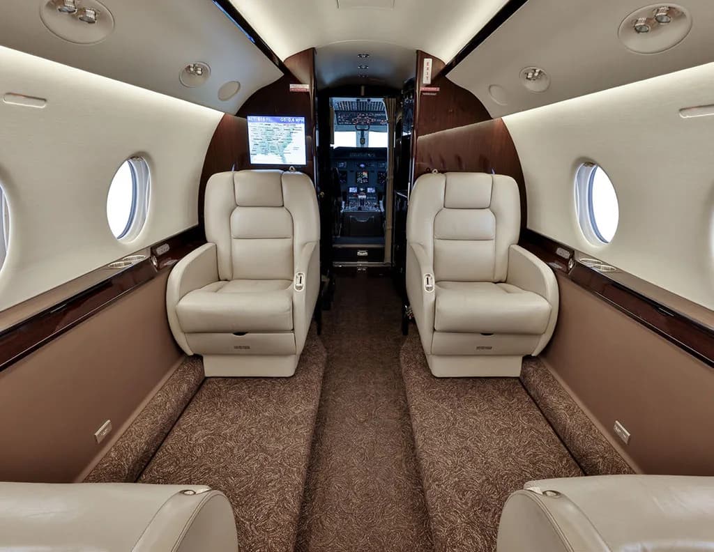 The private jet cabin interior with beige leather seats and a view into the cockpit area.