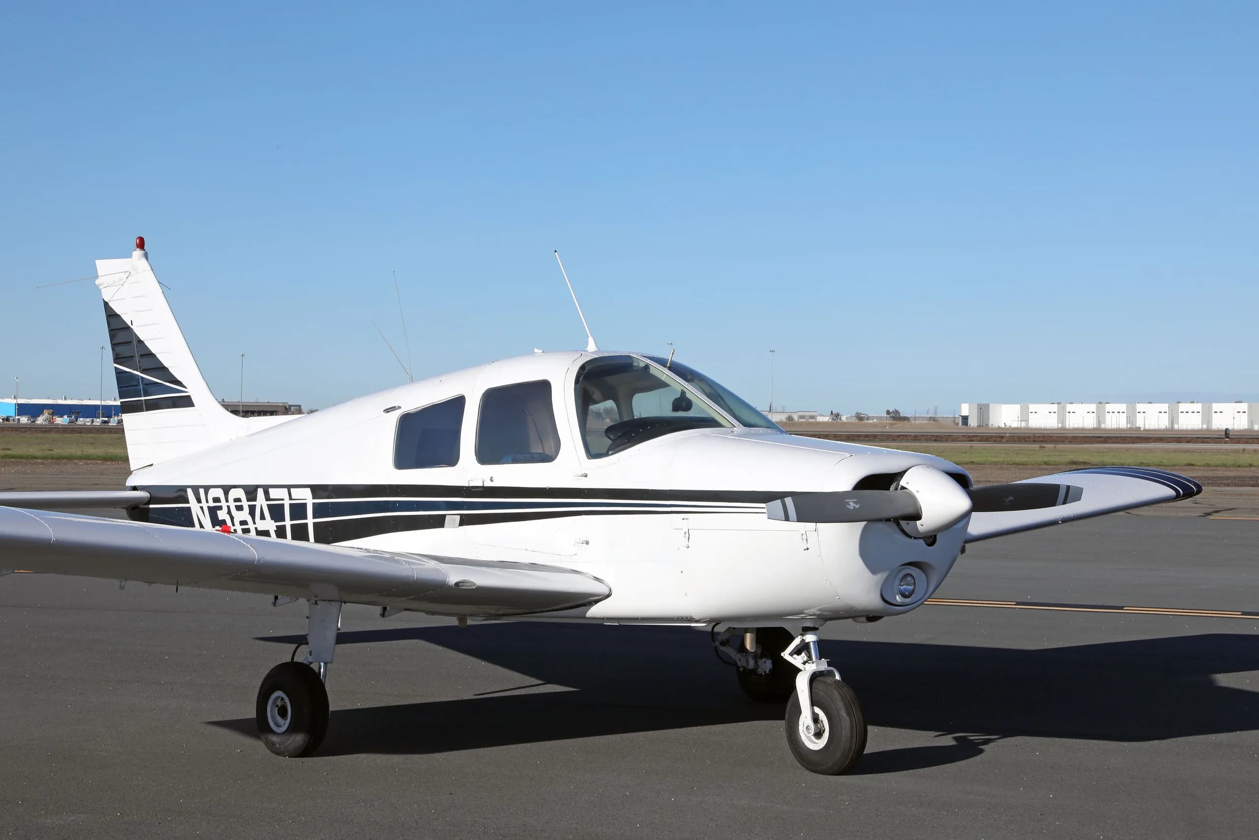 A front three-quarter view of the white Piper Cherokee N38477 with blue and black stripes on an airfield.