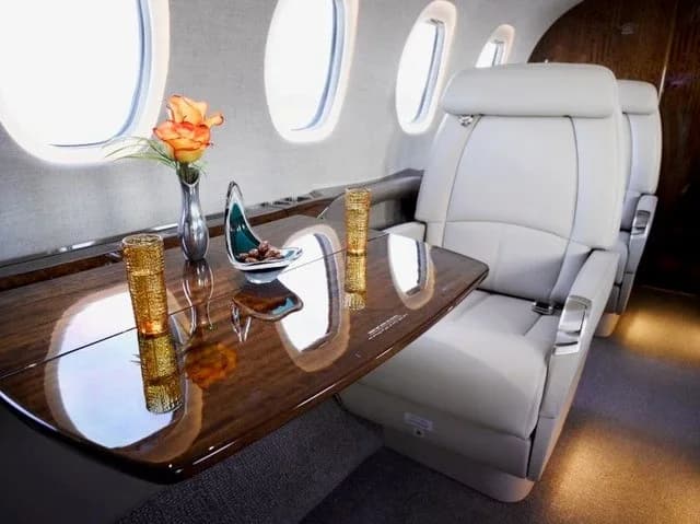 A close-up of a light grey leather seat and a polished wooden table with a vase of flowers and drinks in the aircraft cabin.
