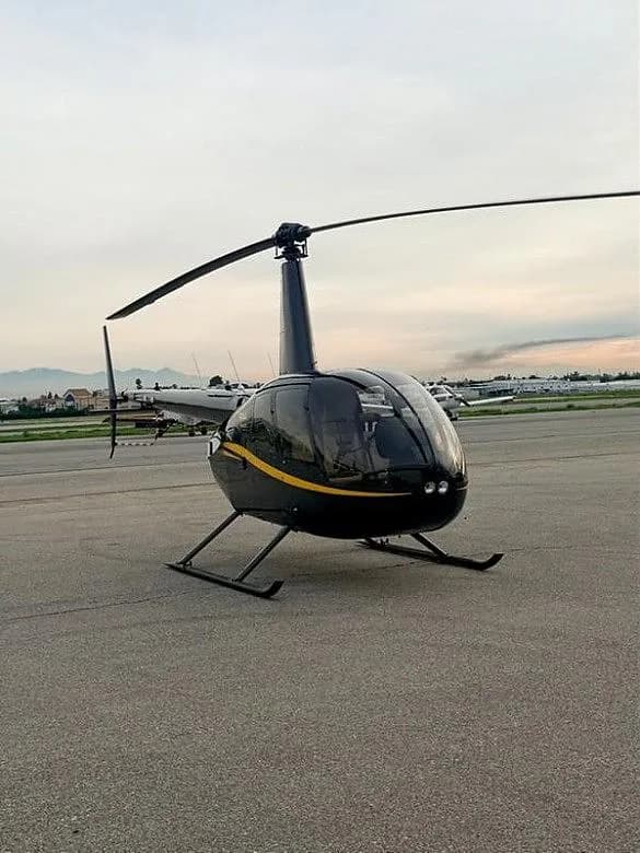 A black Robinson R44 helicopter with a yellow stripe, parked on an airfield with mountains in the background at dusk.