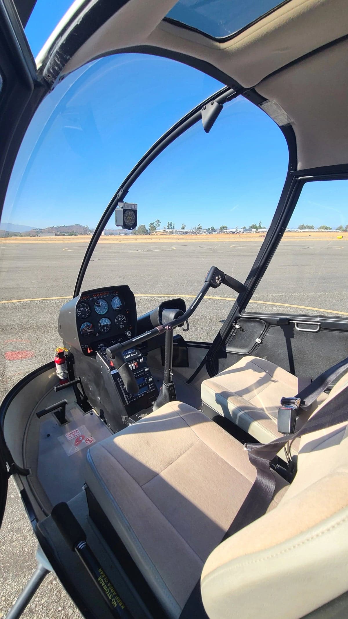The cockpit of a Robinson R22 helicopter, showing the instrument panel, controls, and tan seats, with the runway visible through the windshield.