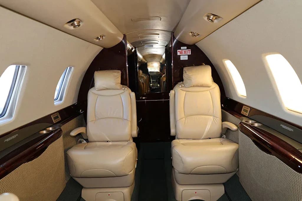 A view down the aisle of a private jet cabin, showing two rows of beige leather seats and dark wood trim.