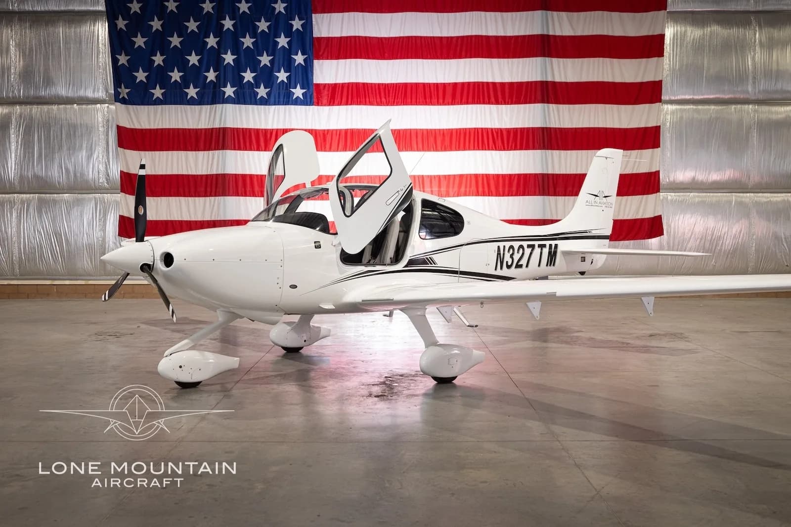 A white Cirrus SR22 aircraft with black stripes and registration N327TM, parked in a hangar with an American flag backdrop, with its gull-wing doors open.