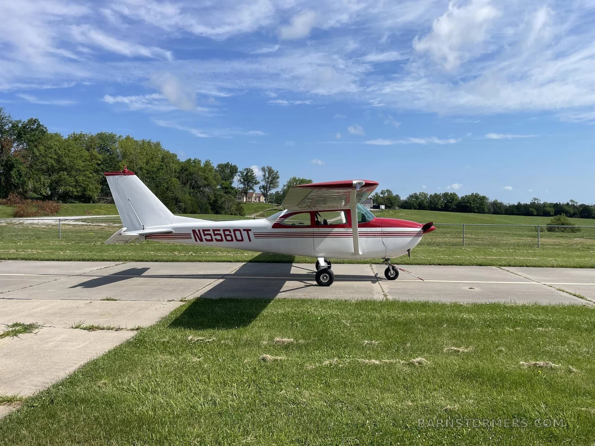 A white and red Cessna N5560T aircraft is parked on a concrete pad with grass and trees in the background under a blue sky.