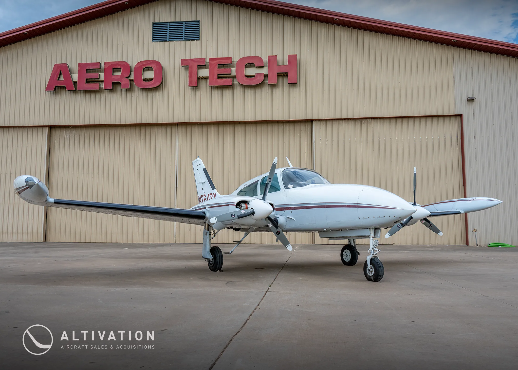 A white twin-engine propeller aircraft with maroon stripes, tail number N2642Y, parked in front of an "AERO TECH" hangar.