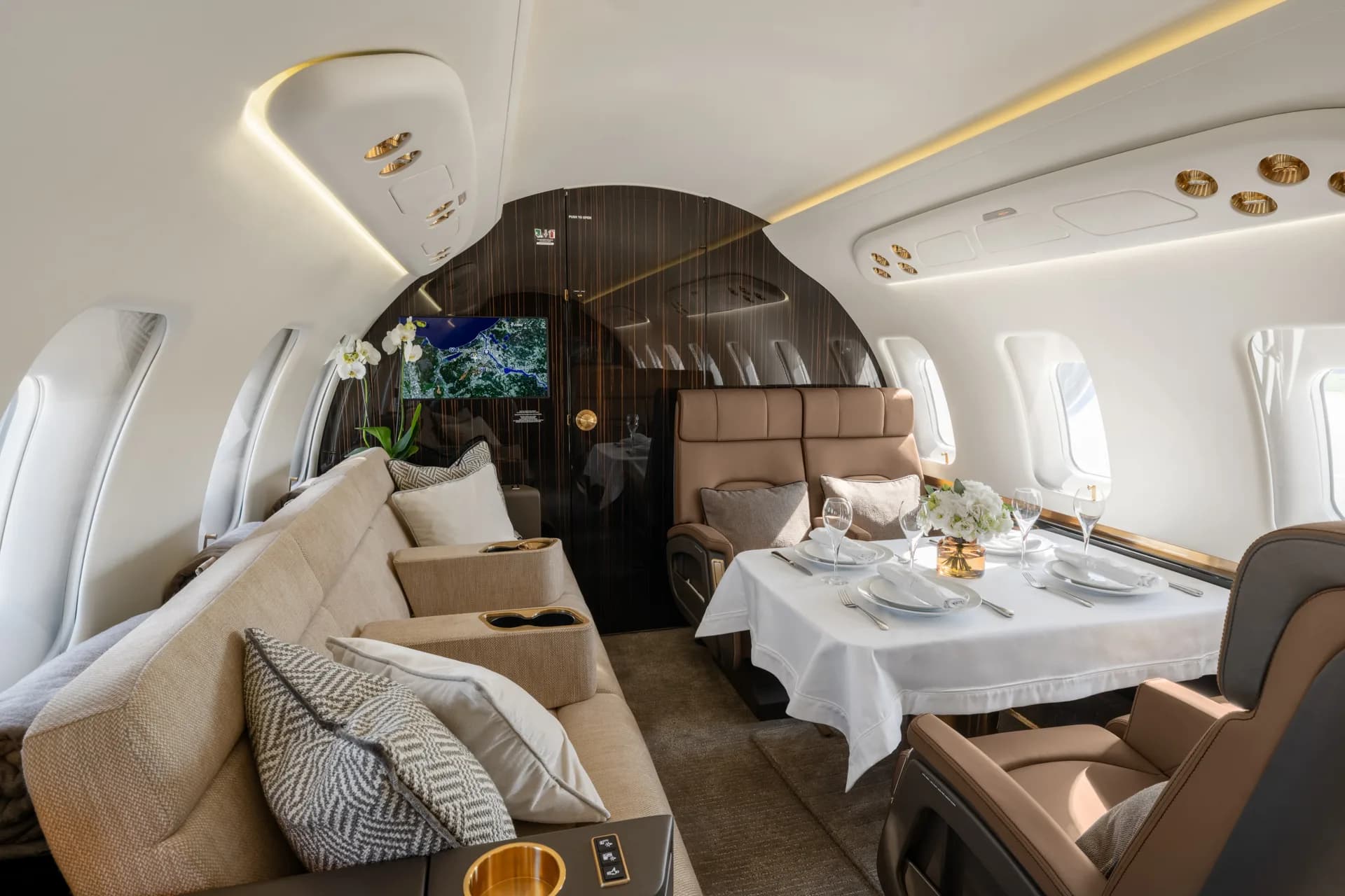 A luxurious aircraft cabin interior featuring a dining table set for four, a beige sofa, and brown leather seats.