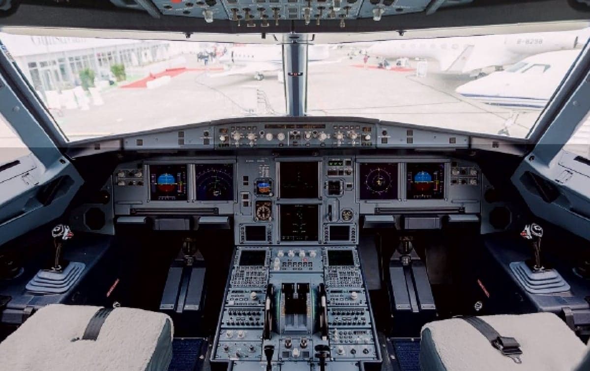 The cockpit of an Airbus aircraft with its instrument panel visible, looking out onto an airfield with other planes.