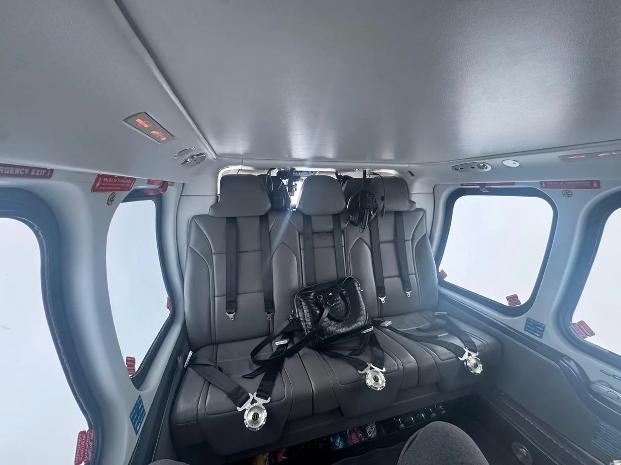 The interior cabin of a helicopter shows three grey leather passenger seats with seatbelts, and a black bag resting on one seat.