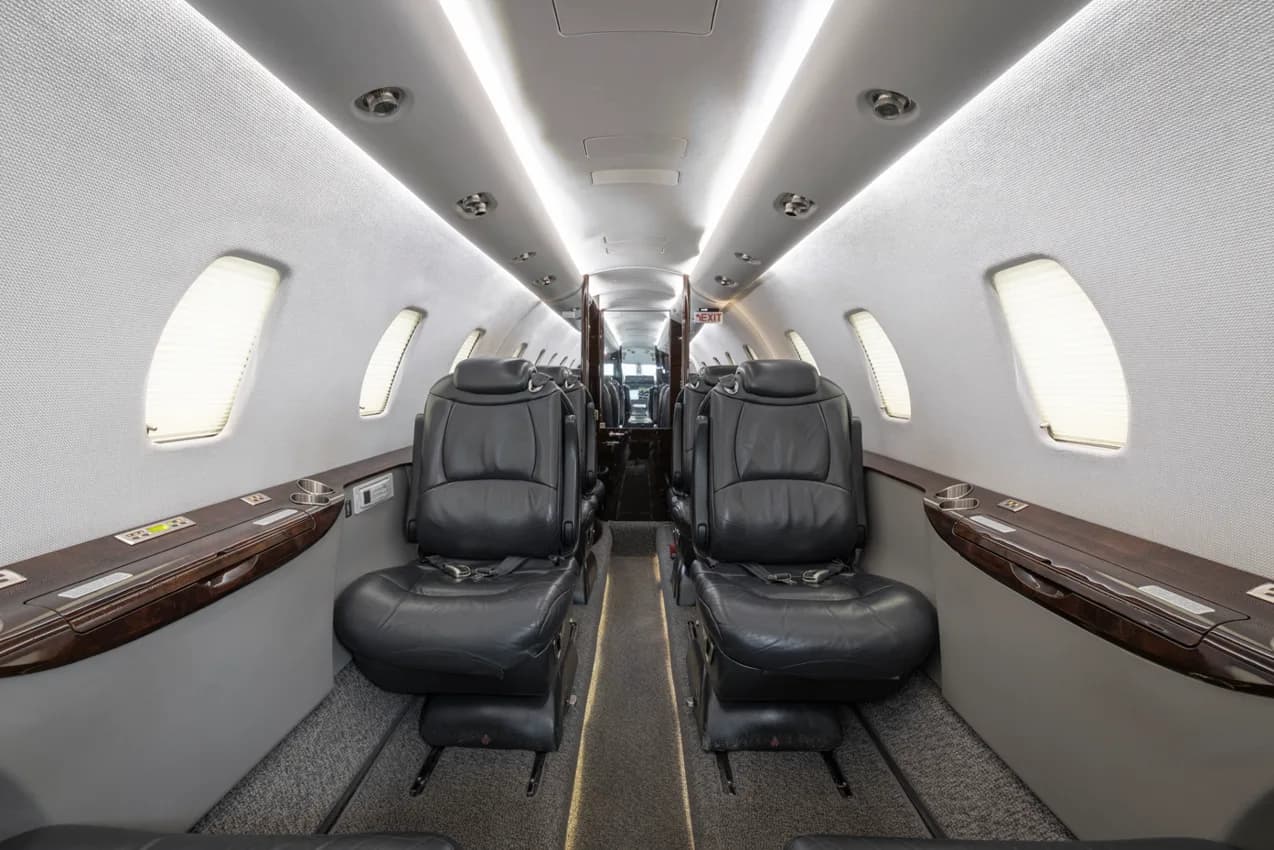 The interior cabin of a business jet, showing rows of black leather seats and overhead lighting.