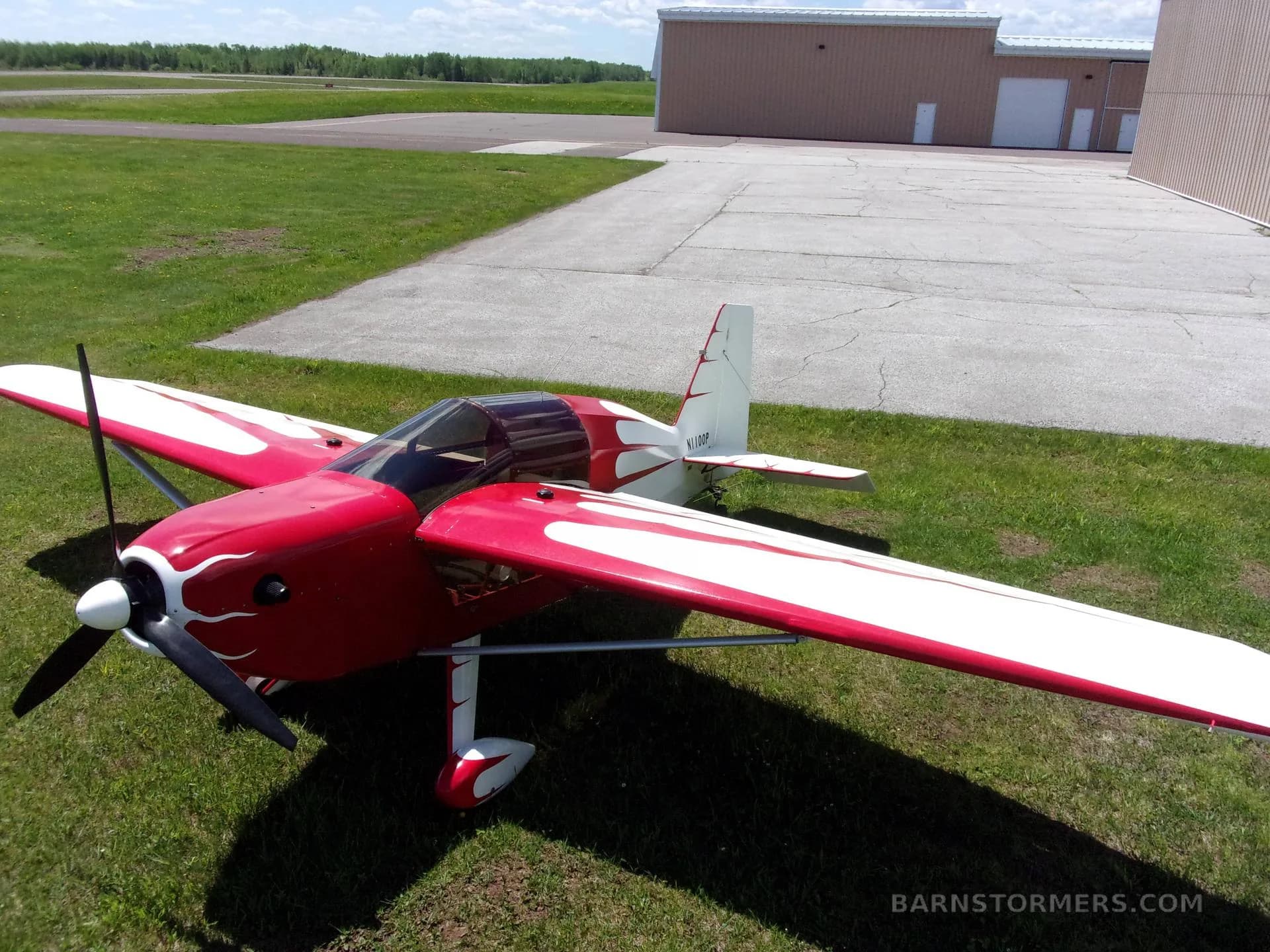A red and white single-engine aircraft with a flame-like paint scheme, parked on grass with hangars in the background.