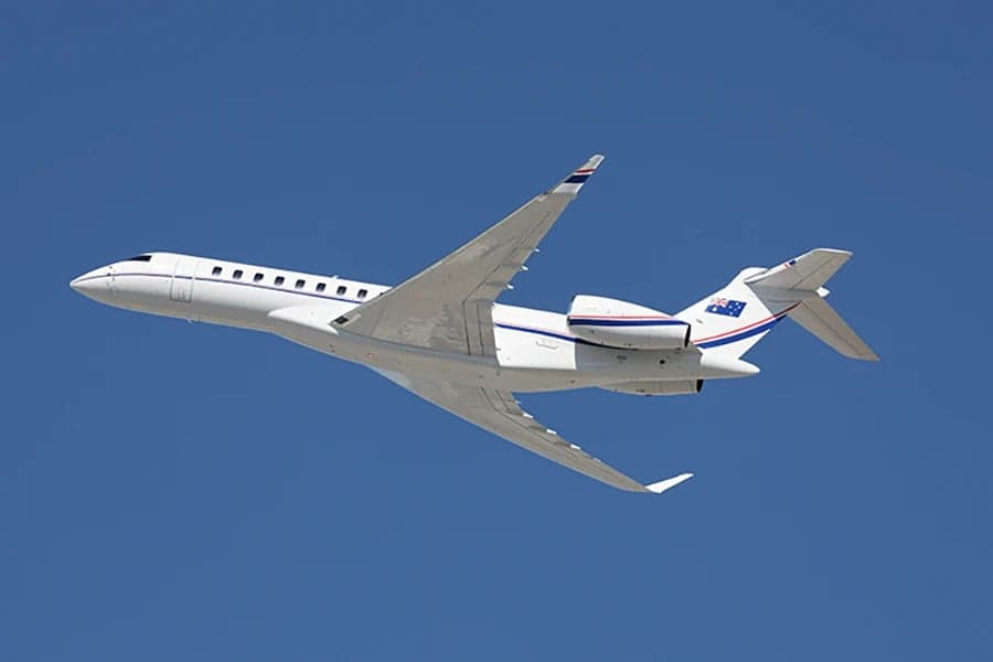 A white Bombardier Global Express business jet in flight against a clear blue sky.