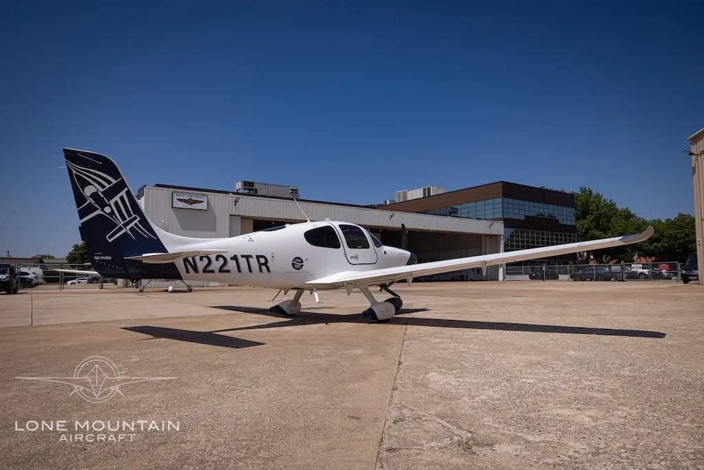 A side view of the white Cirrus SR22 aircraft with a dark blue tail, registration N221TR, parked on an airport tarmac.