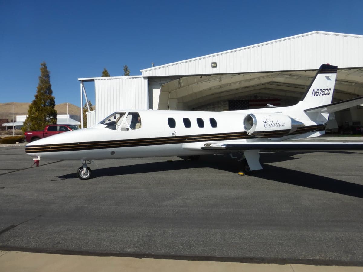 A white Cessna Citation jet with black and gold stripes, registration N676CC, parked on an airfield next to a hangar.