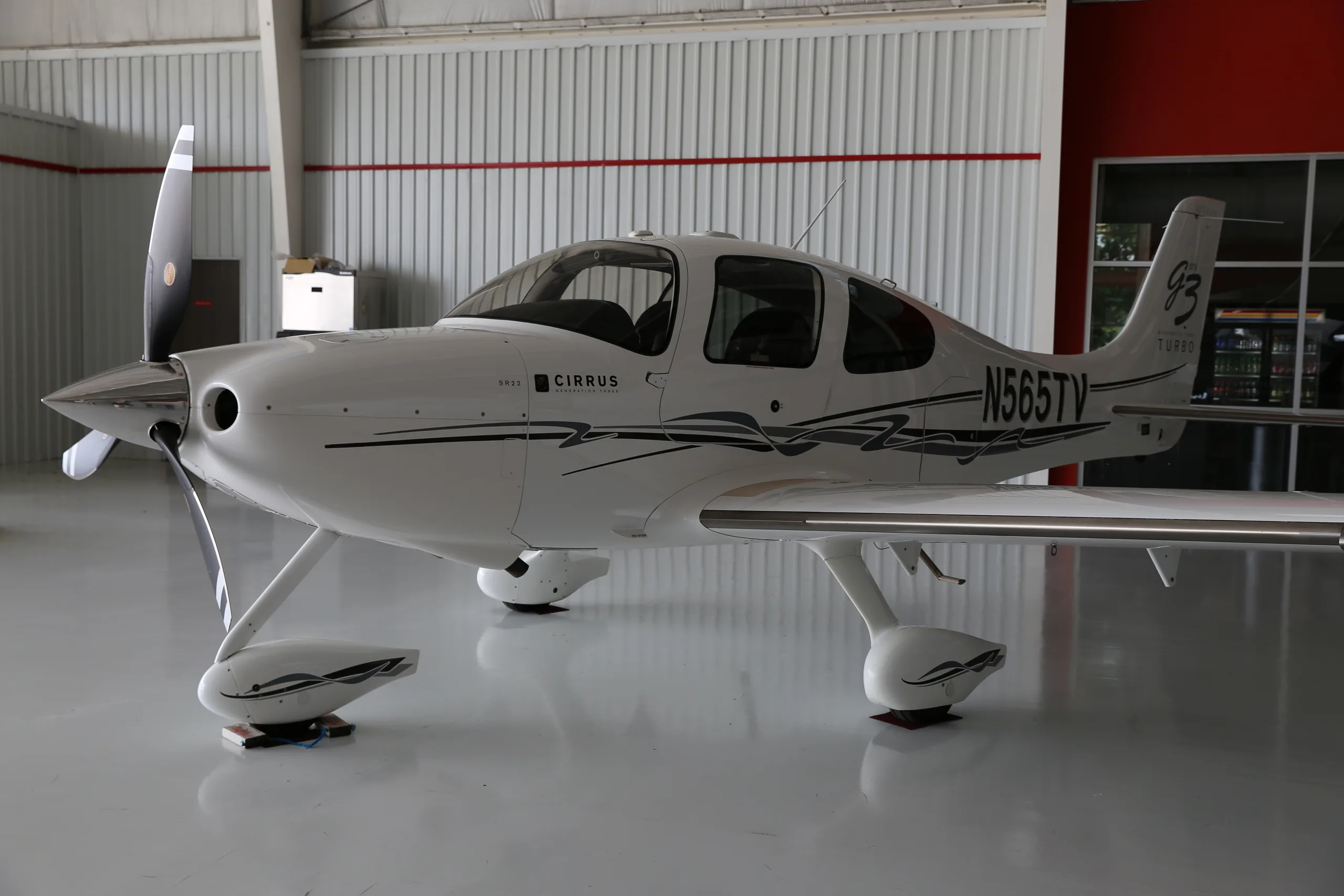 A white Cirrus SR22T aircraft, N565TV, with black and grey stripes, parked inside a hangar.