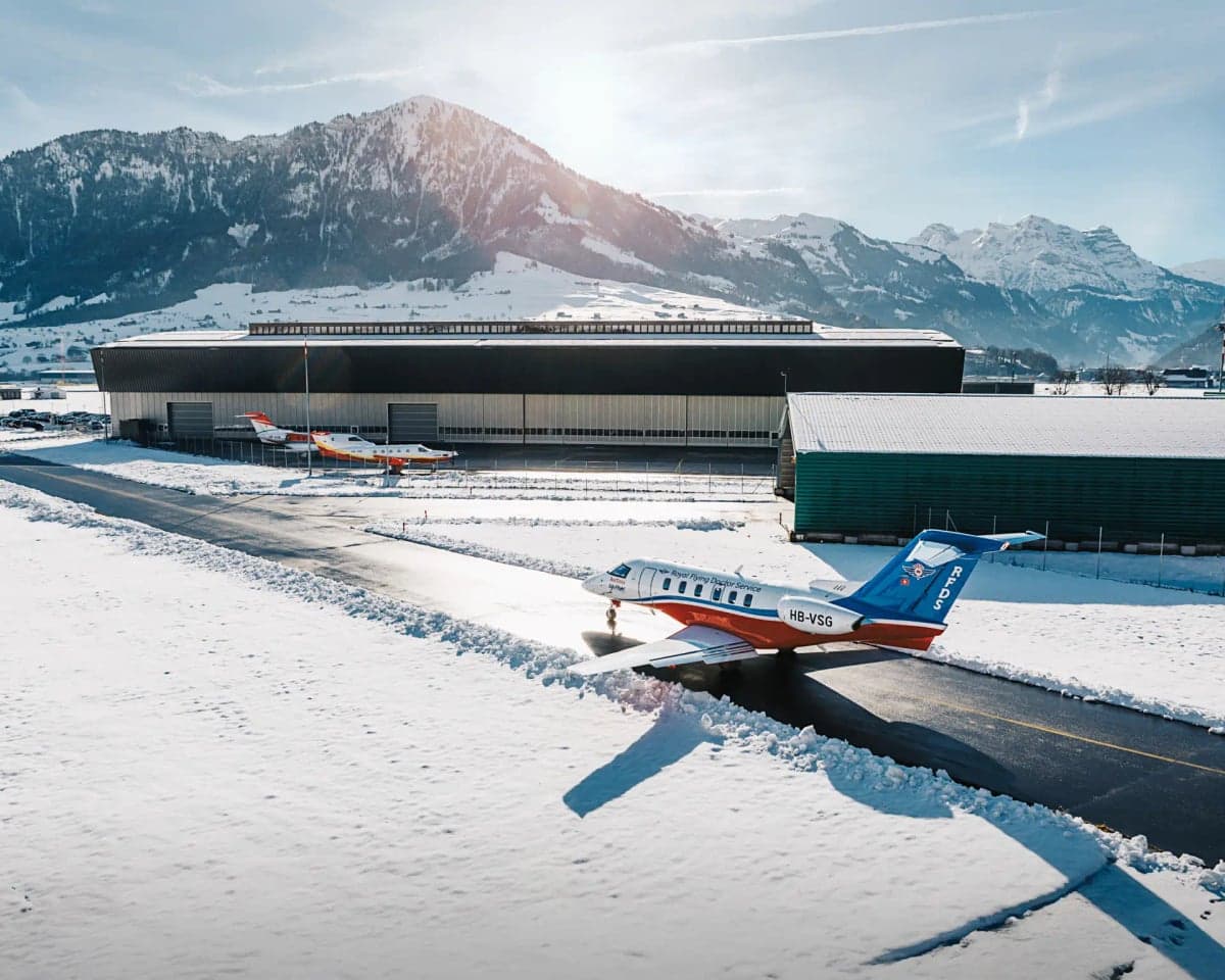 A Pilatus PC-24 aircraft parked on a snowy runway with mountains in the background.