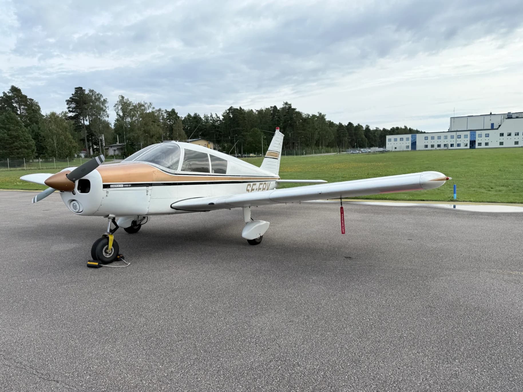 A white and gold Piper Cherokee 140D aircraft is parked on an asphalt apron with trees and a building in the background under a cloudy sky.
