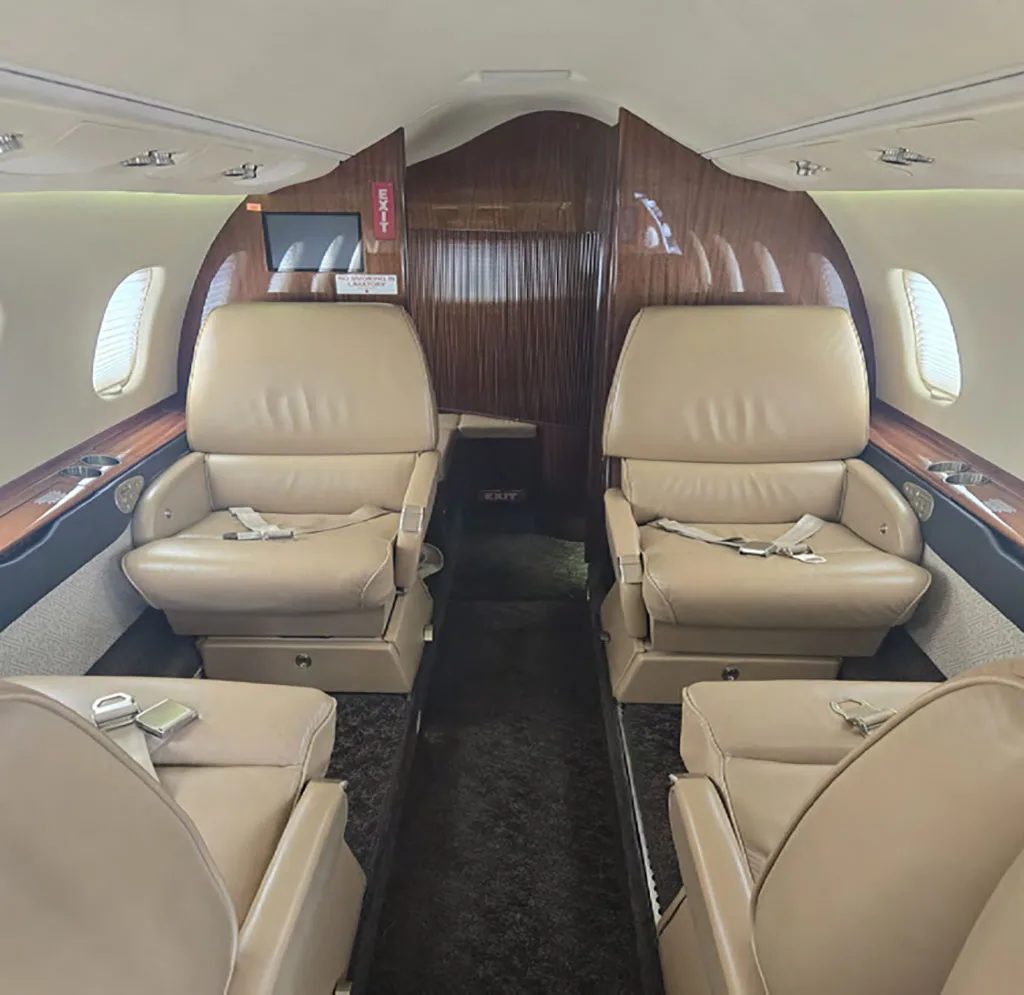 The interior cabin of a business jet, showing tan leather seats and wood paneling, looking towards the rear of the aircraft.