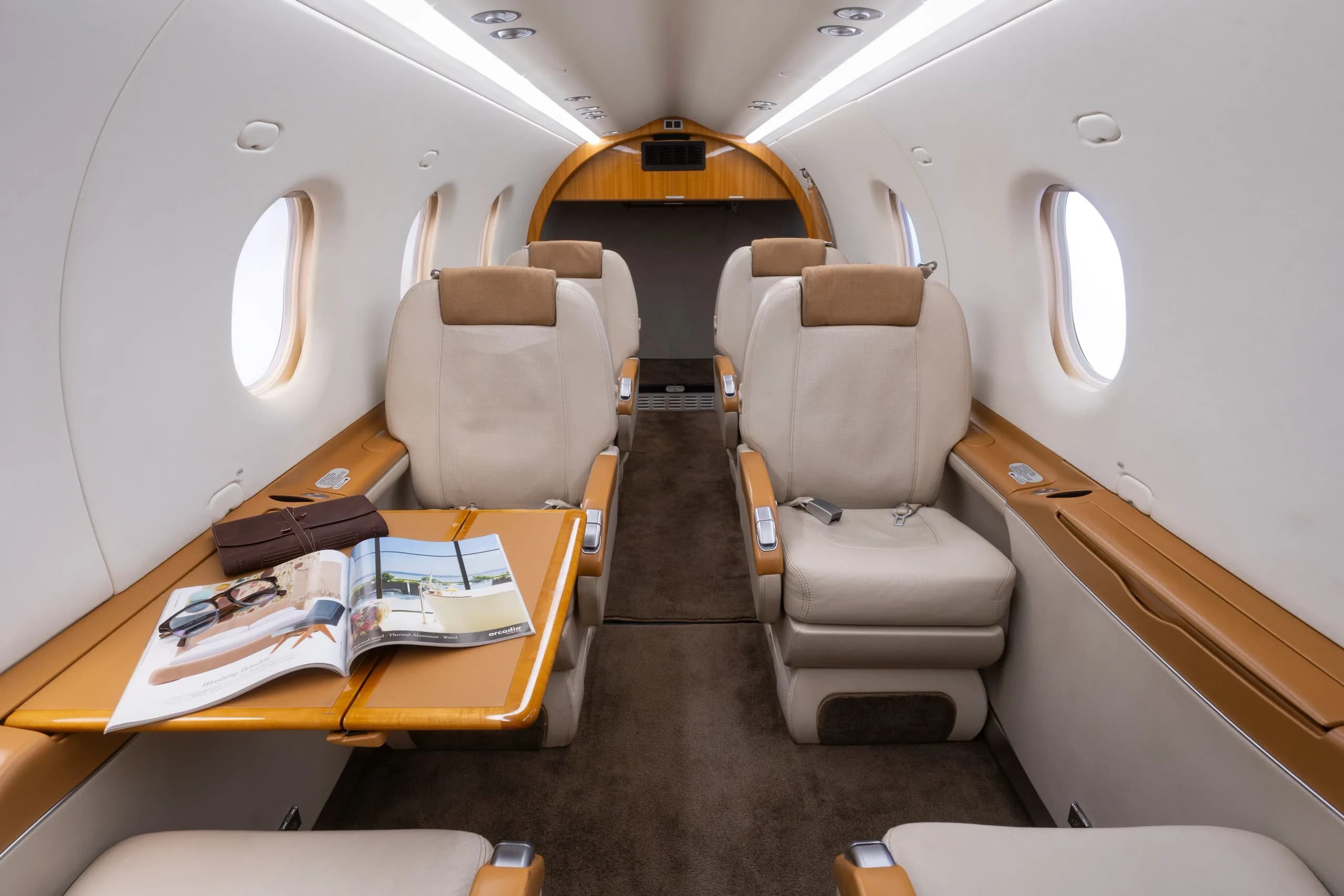 The interior cabin of the Pilatus PC-12, featuring beige leather seats, a wooden table with a magazine, and large windows.
