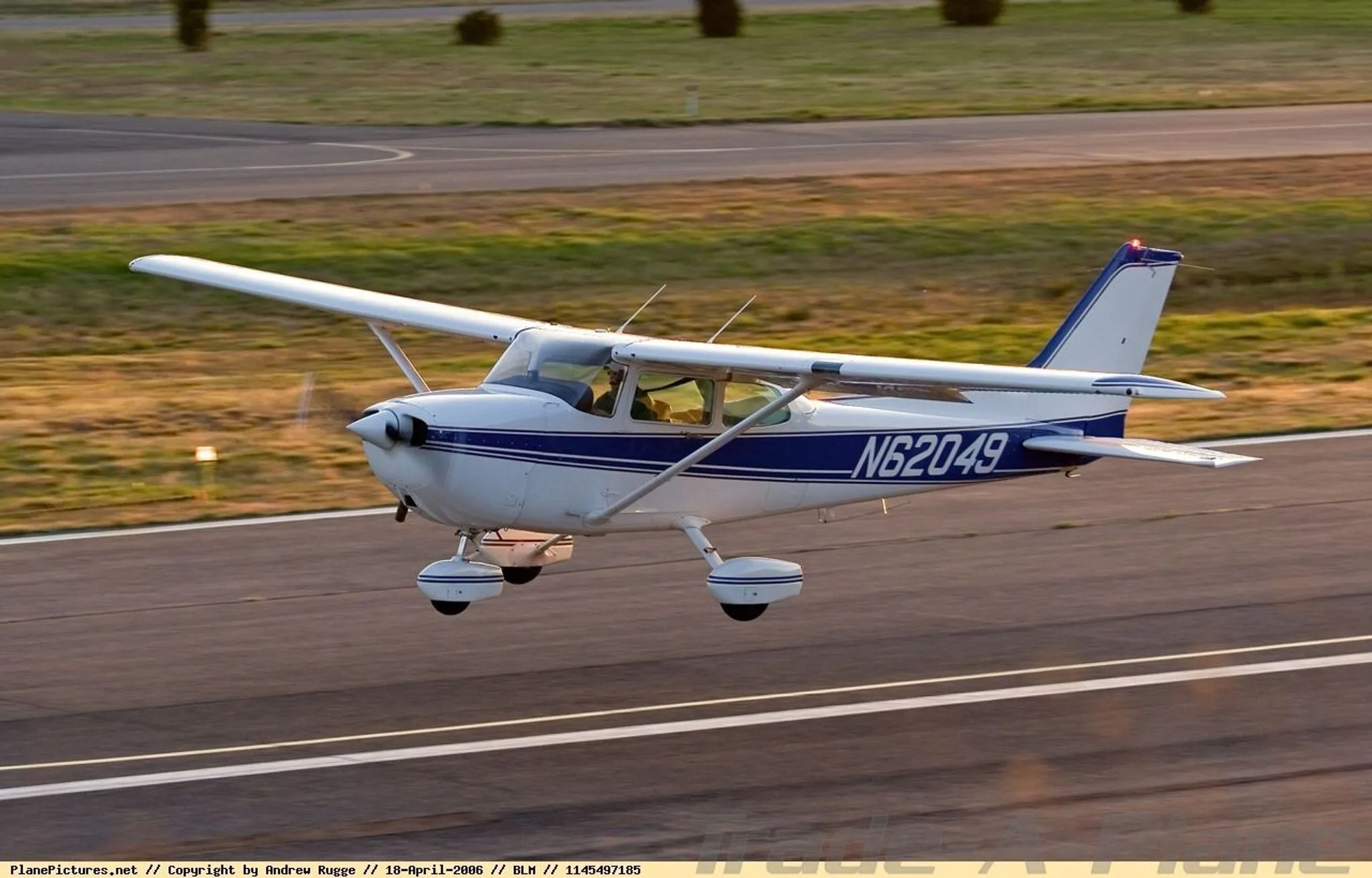 A white Cessna 172 with blue stripes, registration N62049, is captured in flight just above a runway.