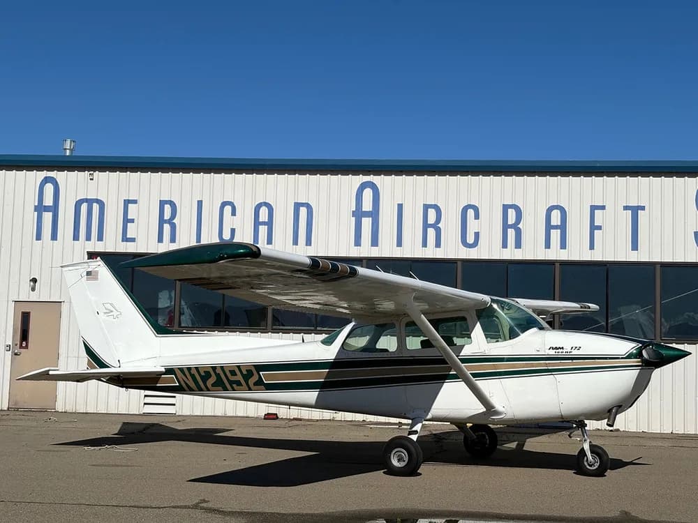 A white Cessna 172 with green and gold stripes parked on an airfield in front of a hangar.