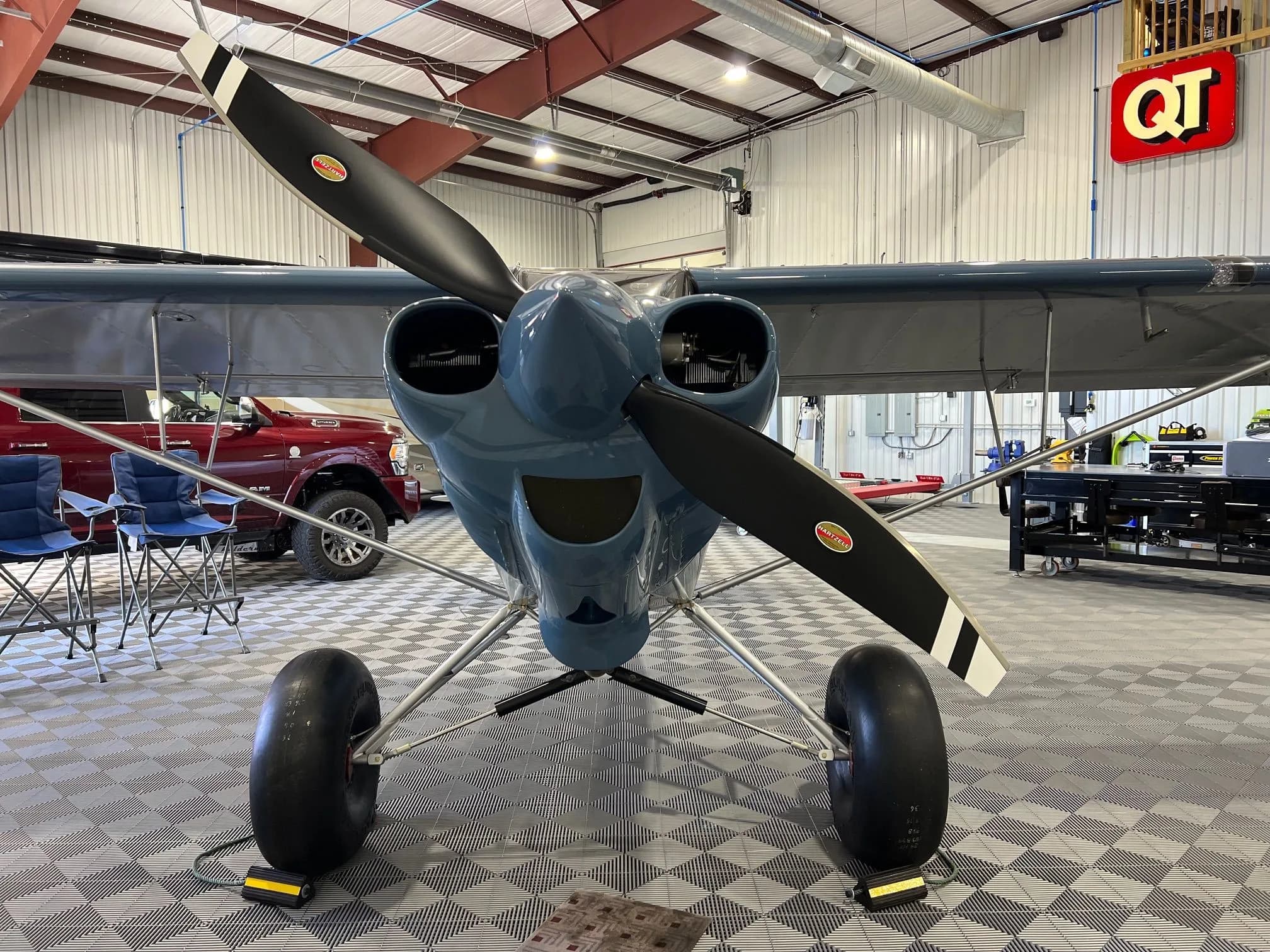 A front view of a blue Carbon Cub FX-3 aircraft with a two-blade propeller, parked inside a hangar on a checkered floor.