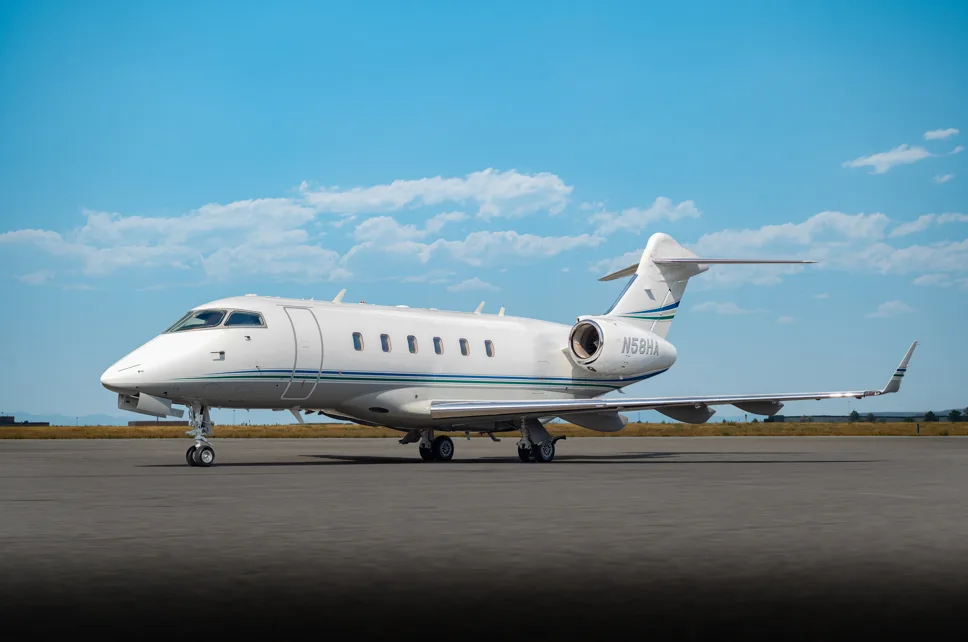 A white Bombardier Challenger business jet with green and blue stripes on the fuselage, parked on a tarmac under a clear blue sky.