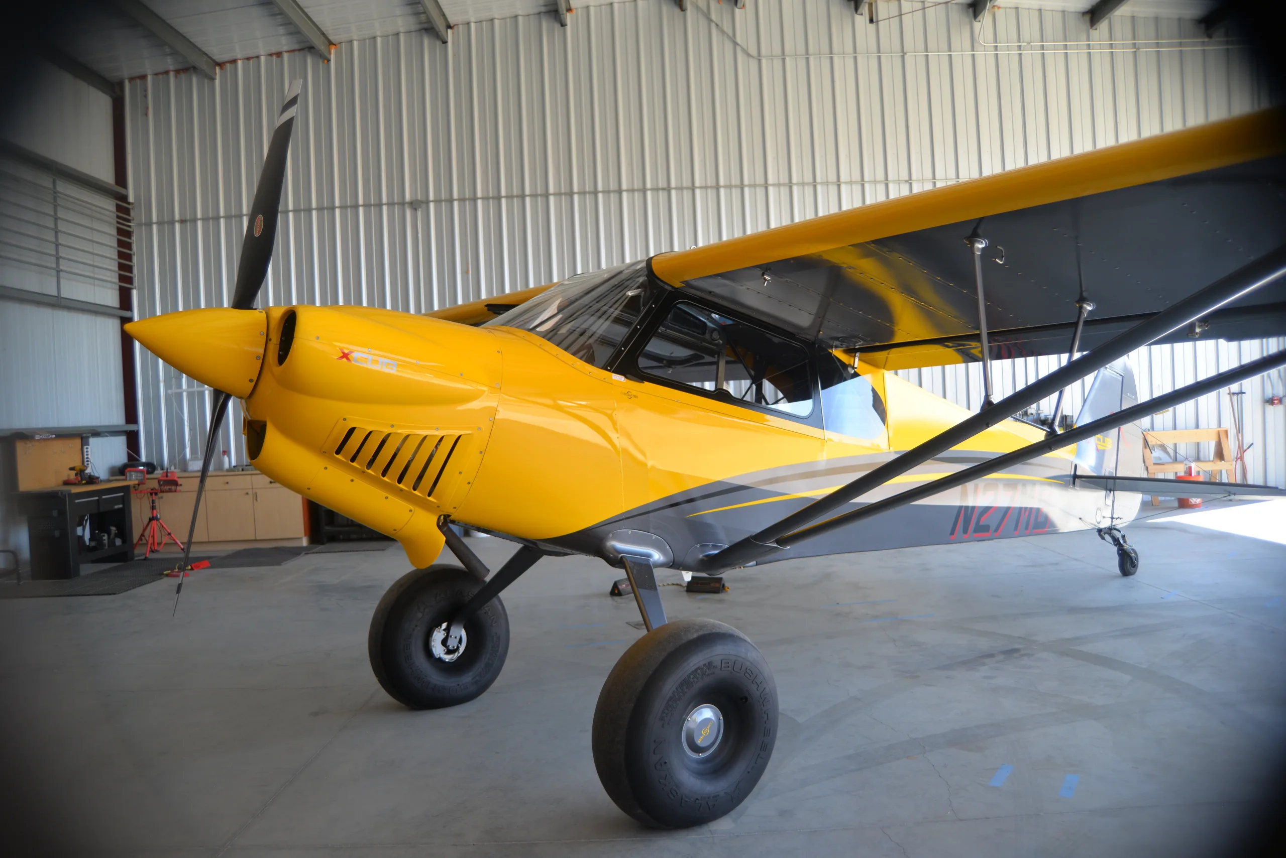 A yellow and gray CubCrafters XCub, registration N27MB, parked inside a hangar.