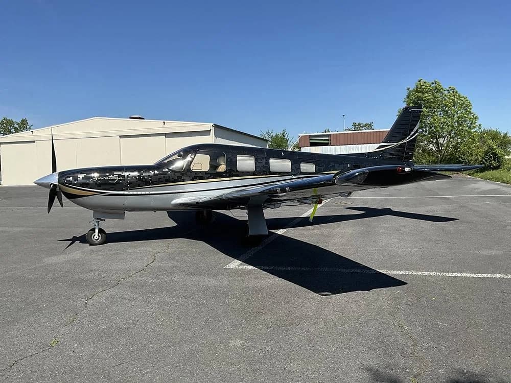 A black and silver Piper Saratoga aircraft with gold accents parked on a tarmac next to a hangar.
