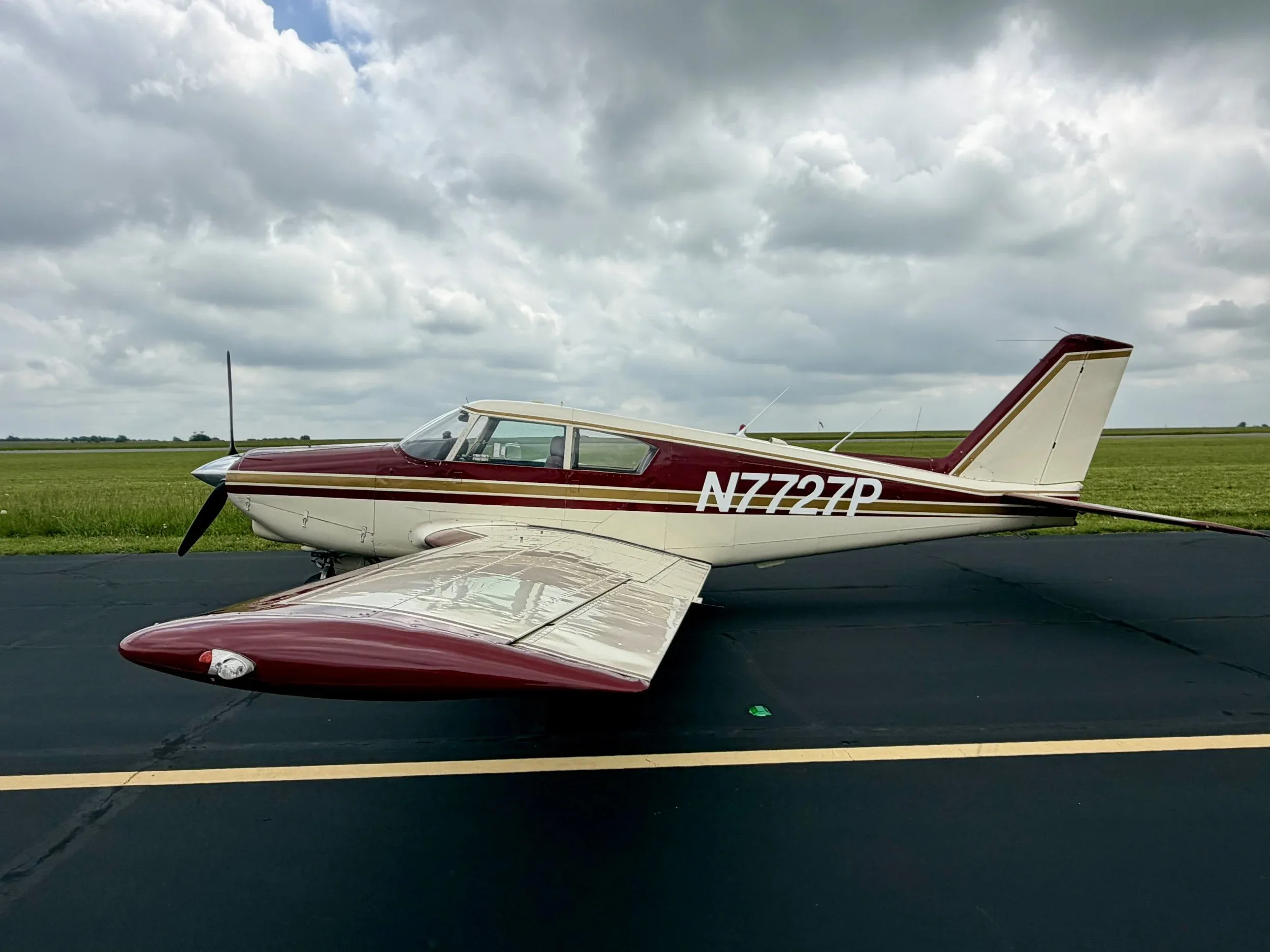 Side view of a maroon and white Piper Comanche N7727P parked on an asphalt runway.