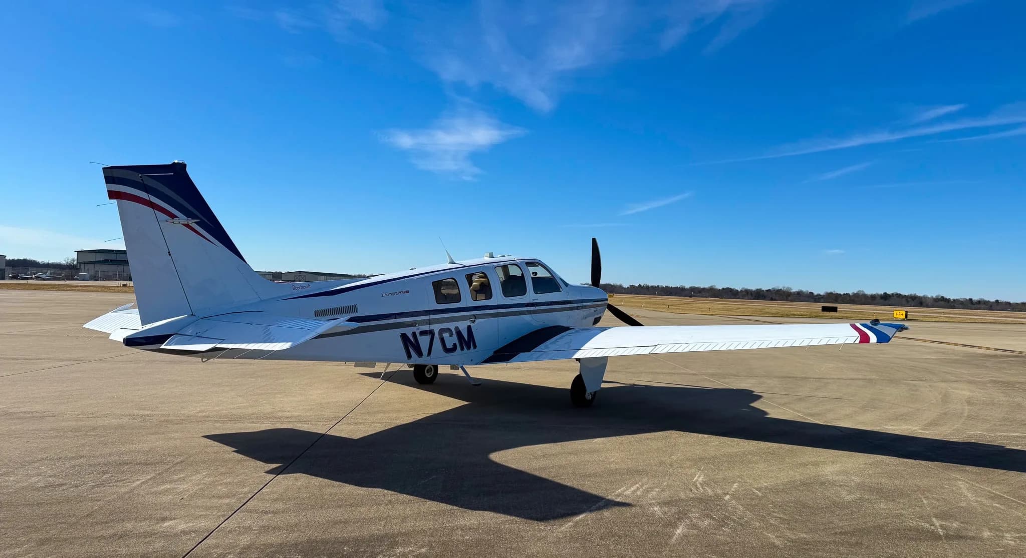 A white Beechcraft Bonanza G36 with blue and grey stripes, registration N7CM, parked on an airfield under a clear blue sky.