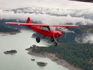 A red Cubcrafters aircraft is flying over a lake and forest.