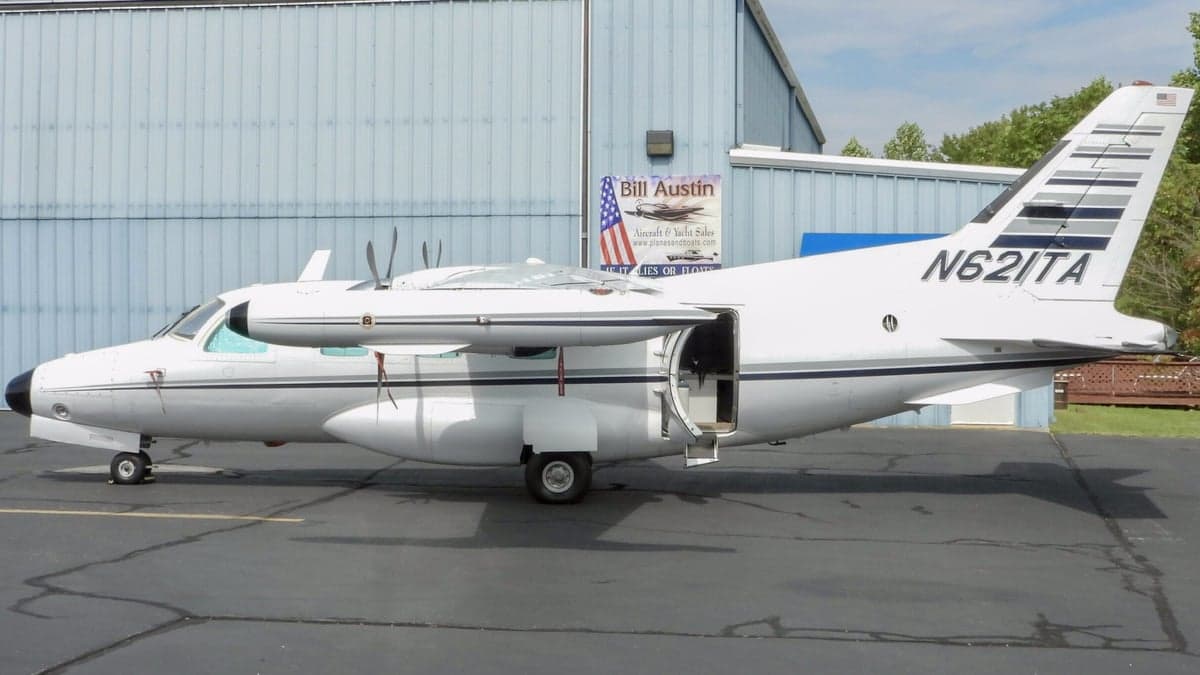 A white Mitsubishi MU-2 aircraft with its passenger door open, parked on an asphalt ramp next to a blue building.
