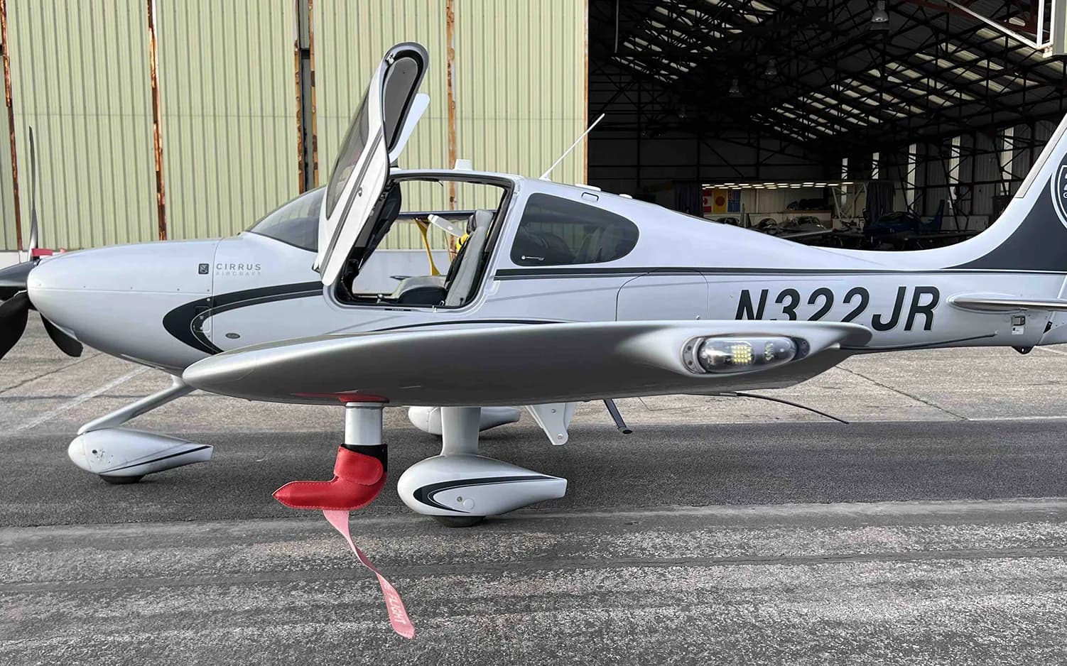 A silver Cirrus SR22 aircraft with registration N322JR and a red "REMOVE BEFORE FLIGHT" tag is parked next to a hangar.