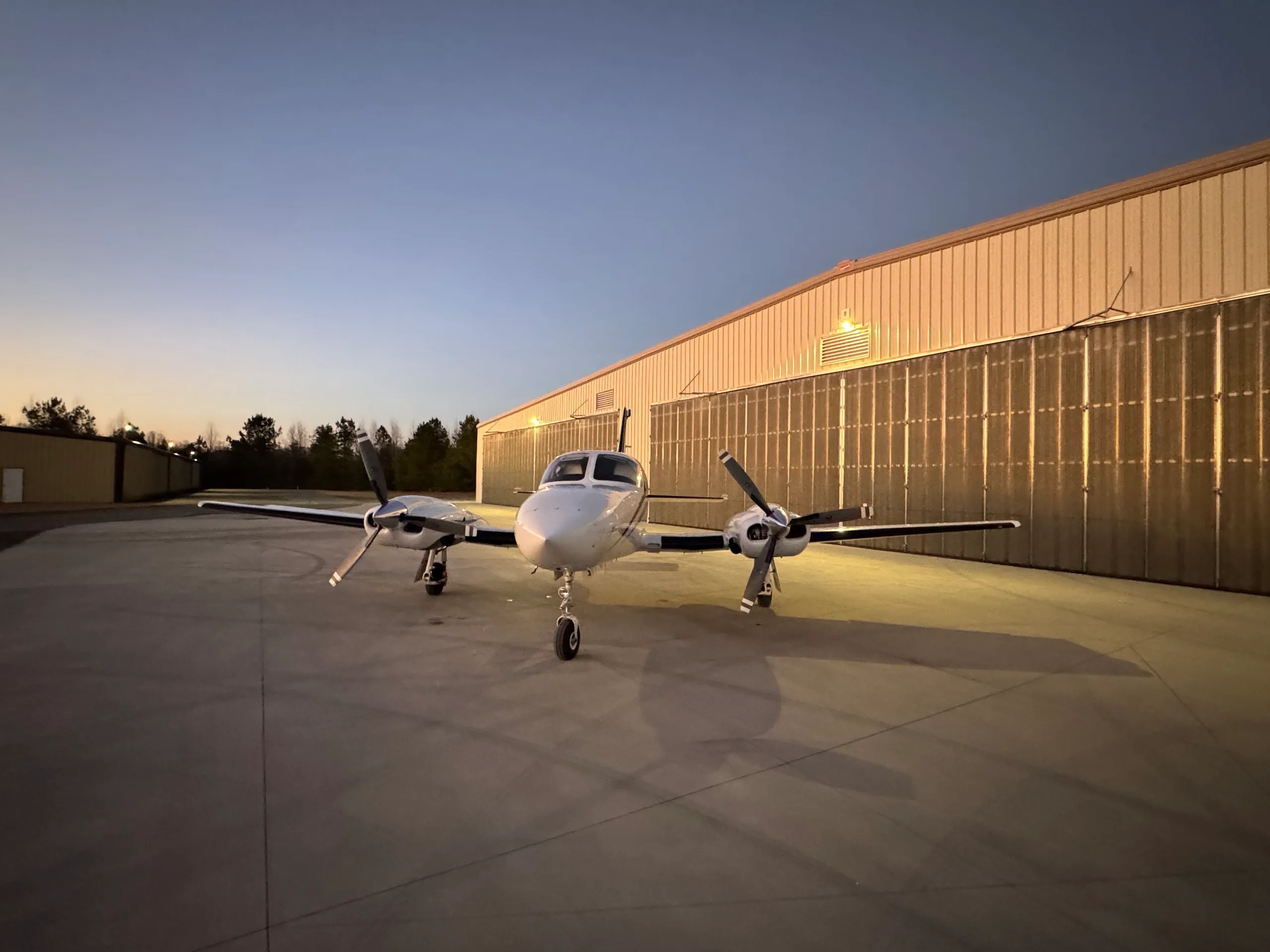 A white twin-engine aircraft with blue stripes, N762CA, viewed head-on while parked in front of a hangar.
