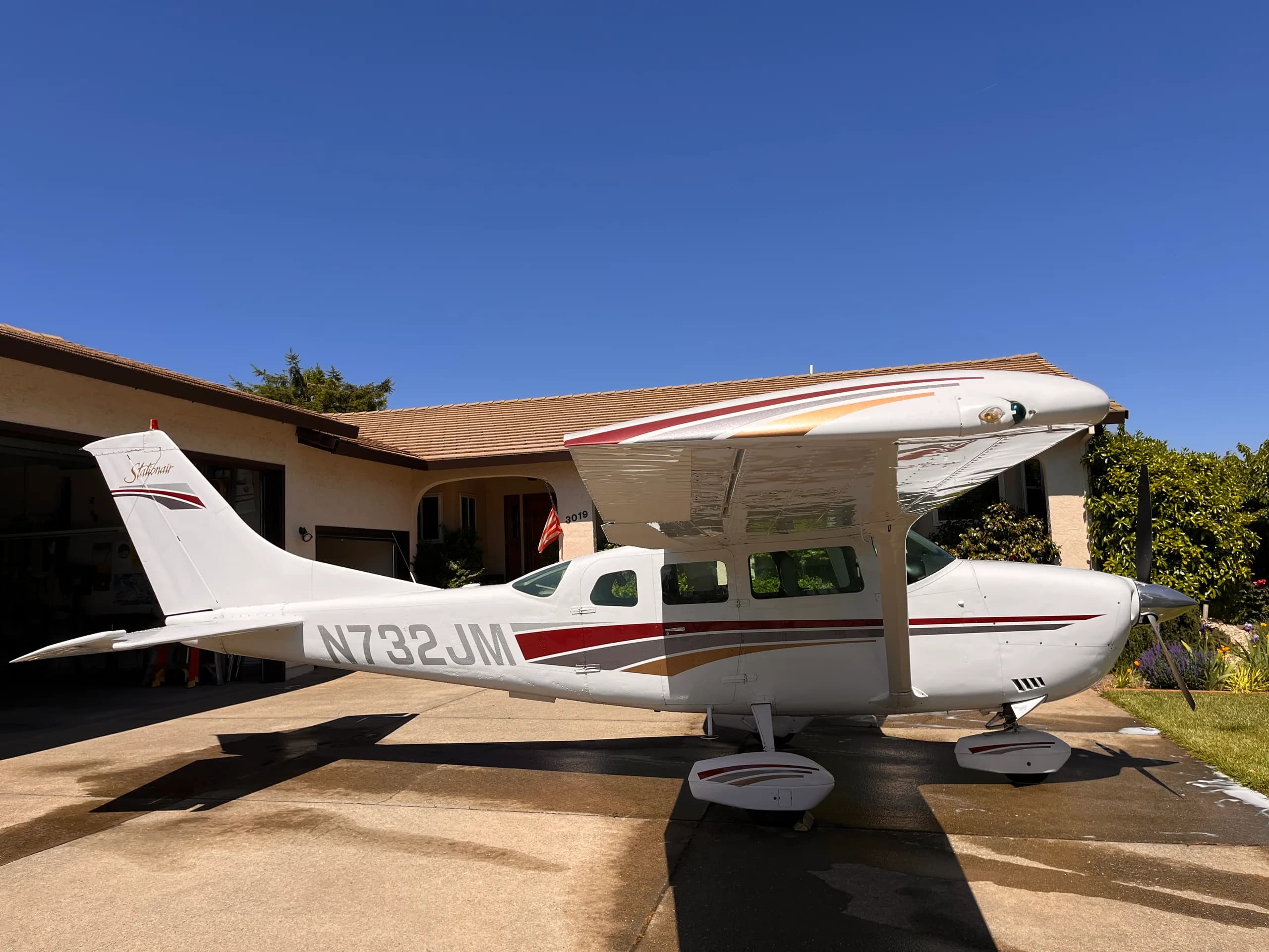 A white Cessna Stationair (N732JM) with red, grey, and gold stripes is parked on a concrete driveway in front of a house on a sunny day.