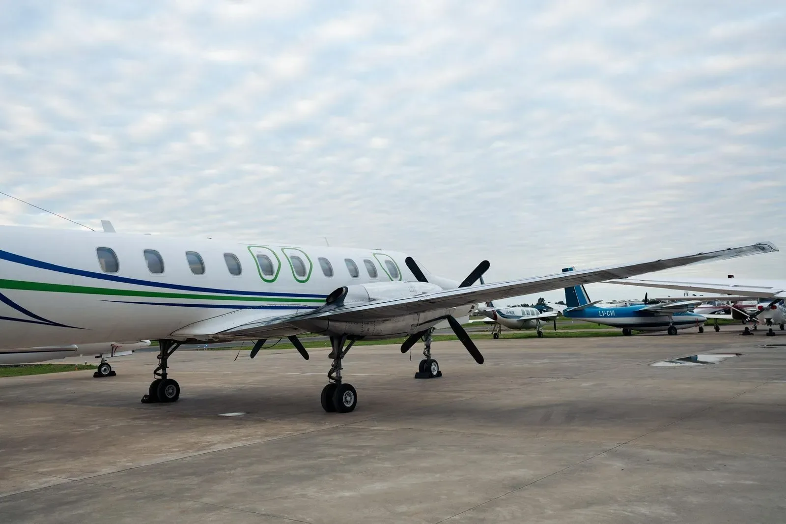 A side view of a white turboprop aircraft with green and blue stripes, parked on an tarmac with other planes in the background.