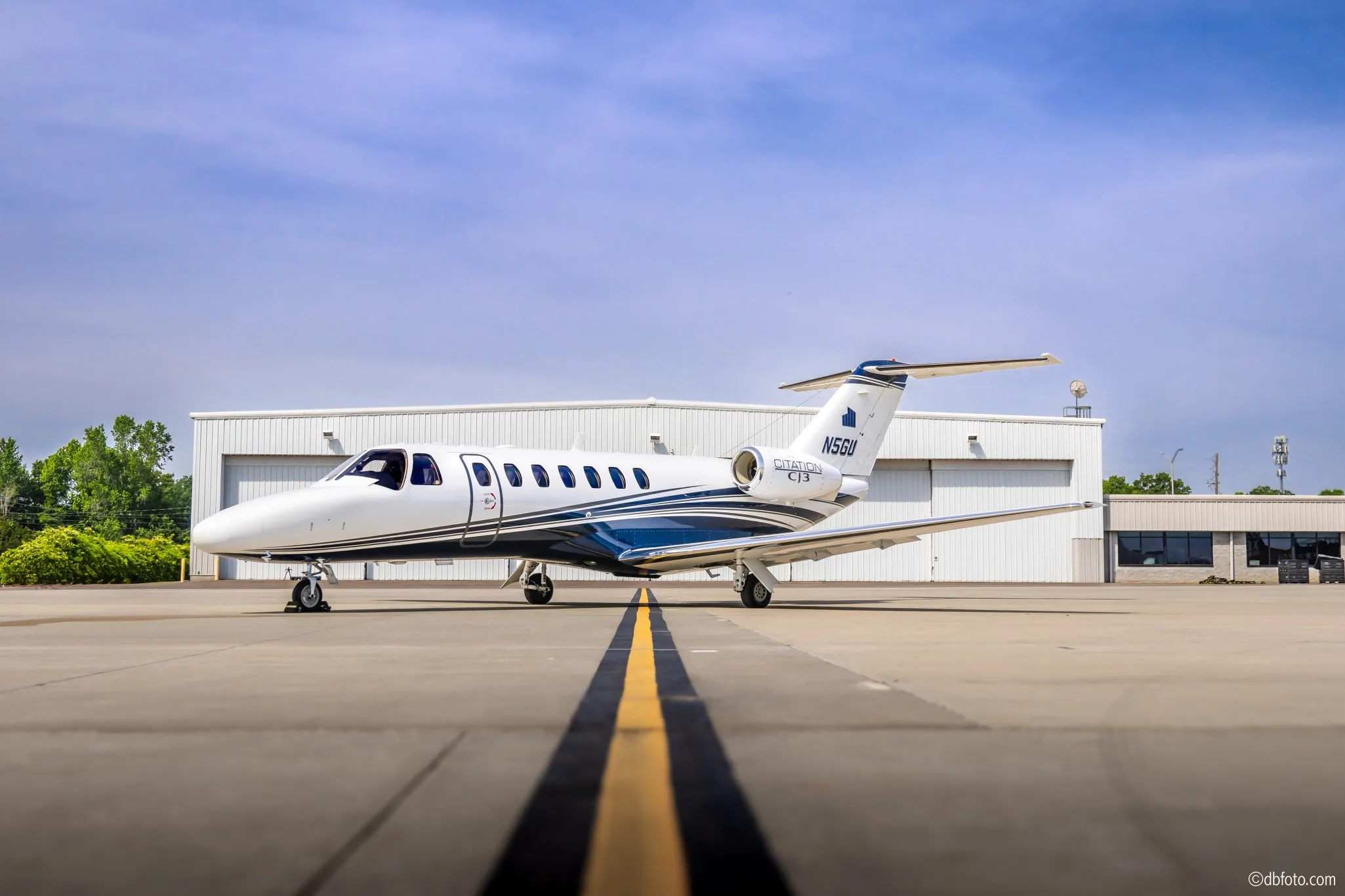 A white Cessna Citation CJ3 business jet with blue and grey stripes, registration N5GU, parked on an airport tarmac in front of hangars.