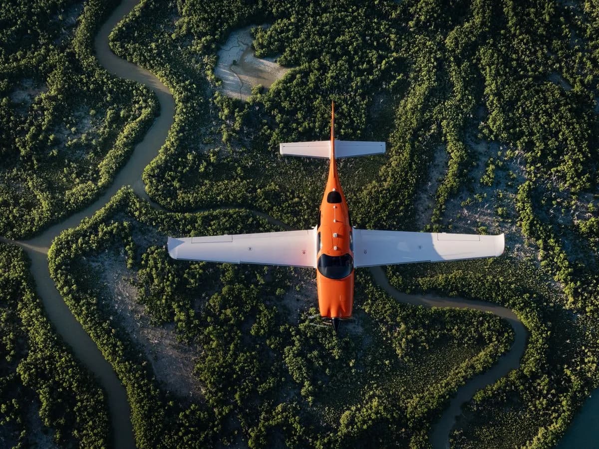 An orange and white single-engine aircraft flies over a winding river through lush green trees, viewed from directly above.