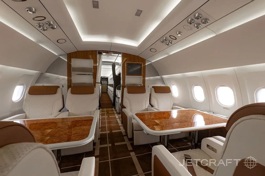 A spacious aircraft cabin interior with cream leather seats, polished wooden tables, and an aisle leading to the rear.