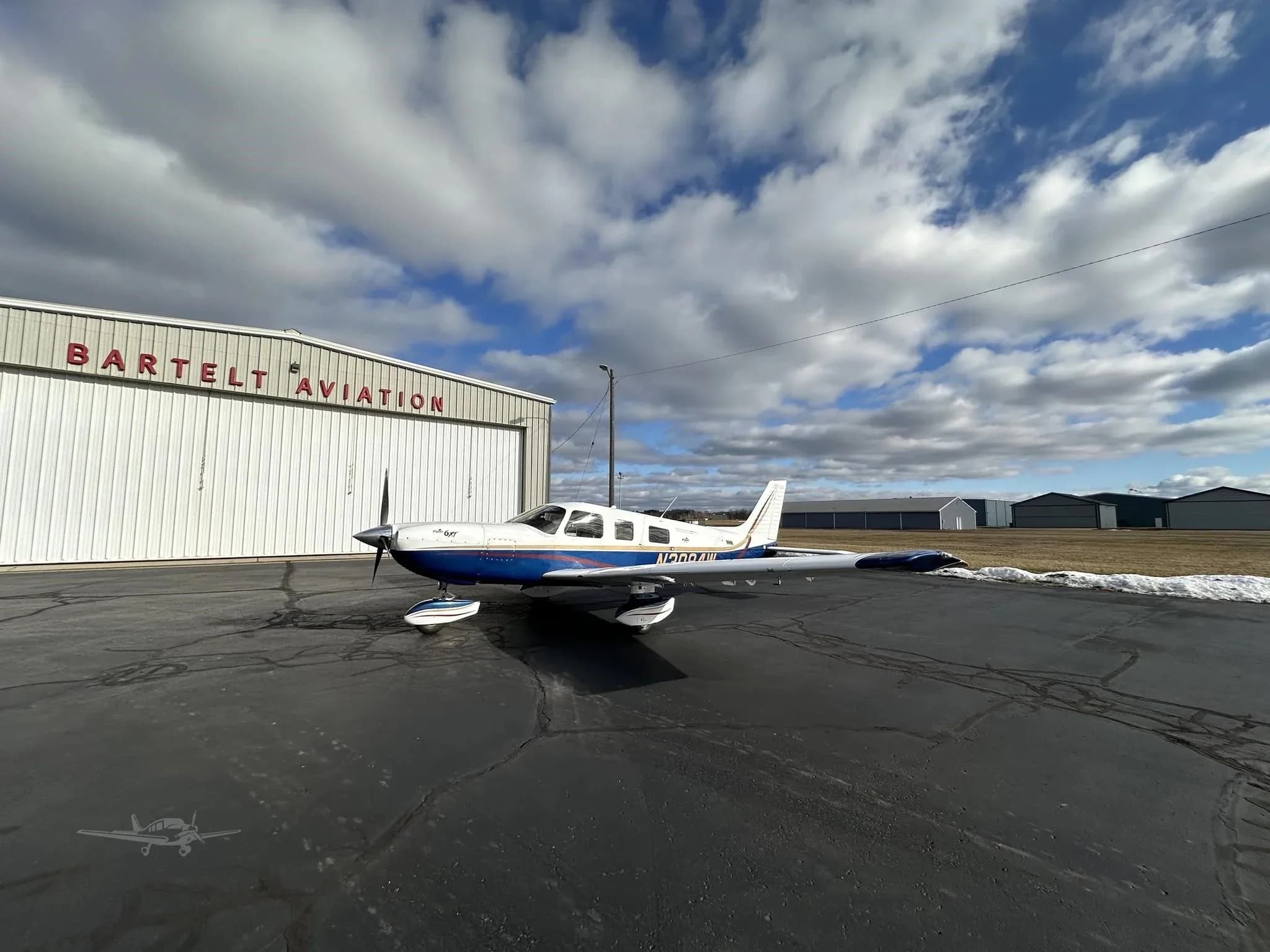 A blue and white Piper Malibu Mirage N3094W parked on an asphalt ramp in front of a Bartelt Aviation hangar under a cloudy sky.